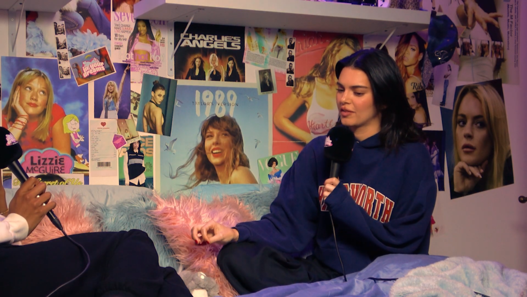 Kendall Jenner sits on a bed in front of a collage of posters of sensible music tastes and films, all from Owen Thiele.